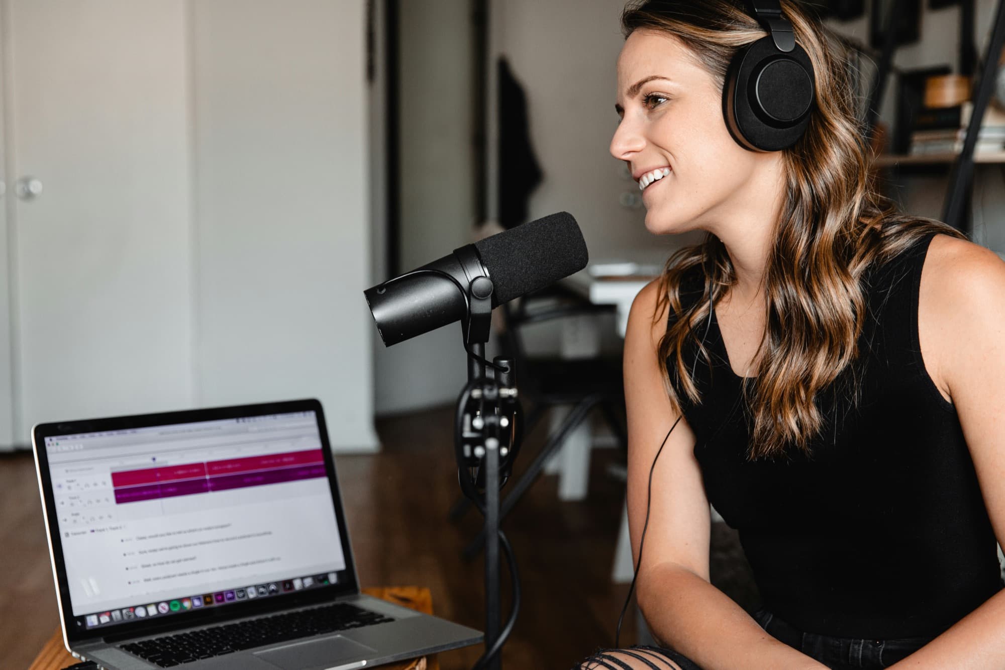 A creator wearing headphones speaking into a microphone beside an open laptop, recording a session in a home studio.